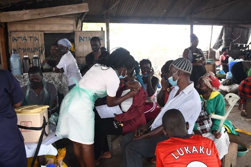 A community health screening session under a covered outdoor space, with families and health workers