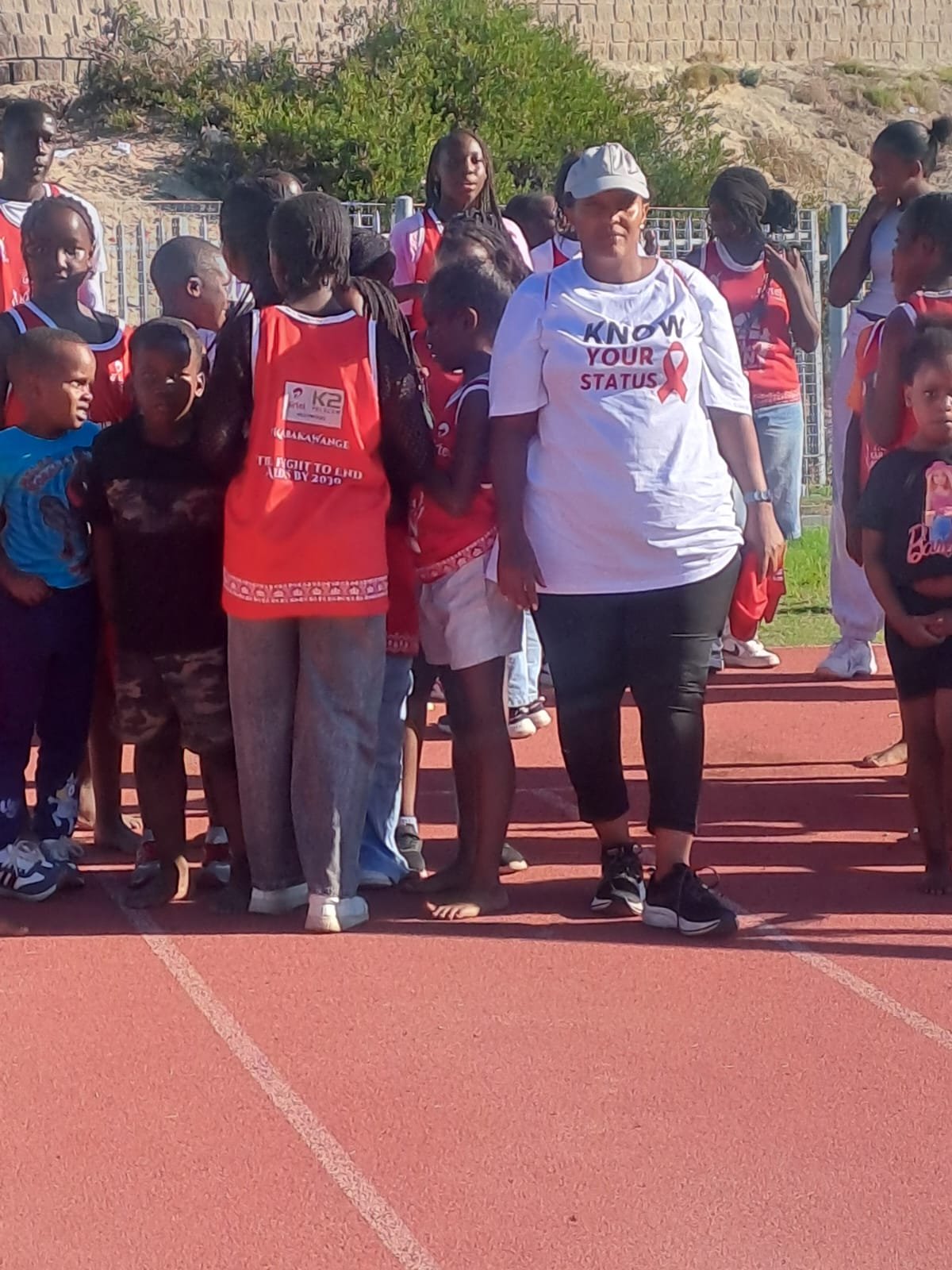 Adolescents on an athletics track alongside a foundation volunteer wearing a 'Know Your Status' t-shirt at a community awareness event